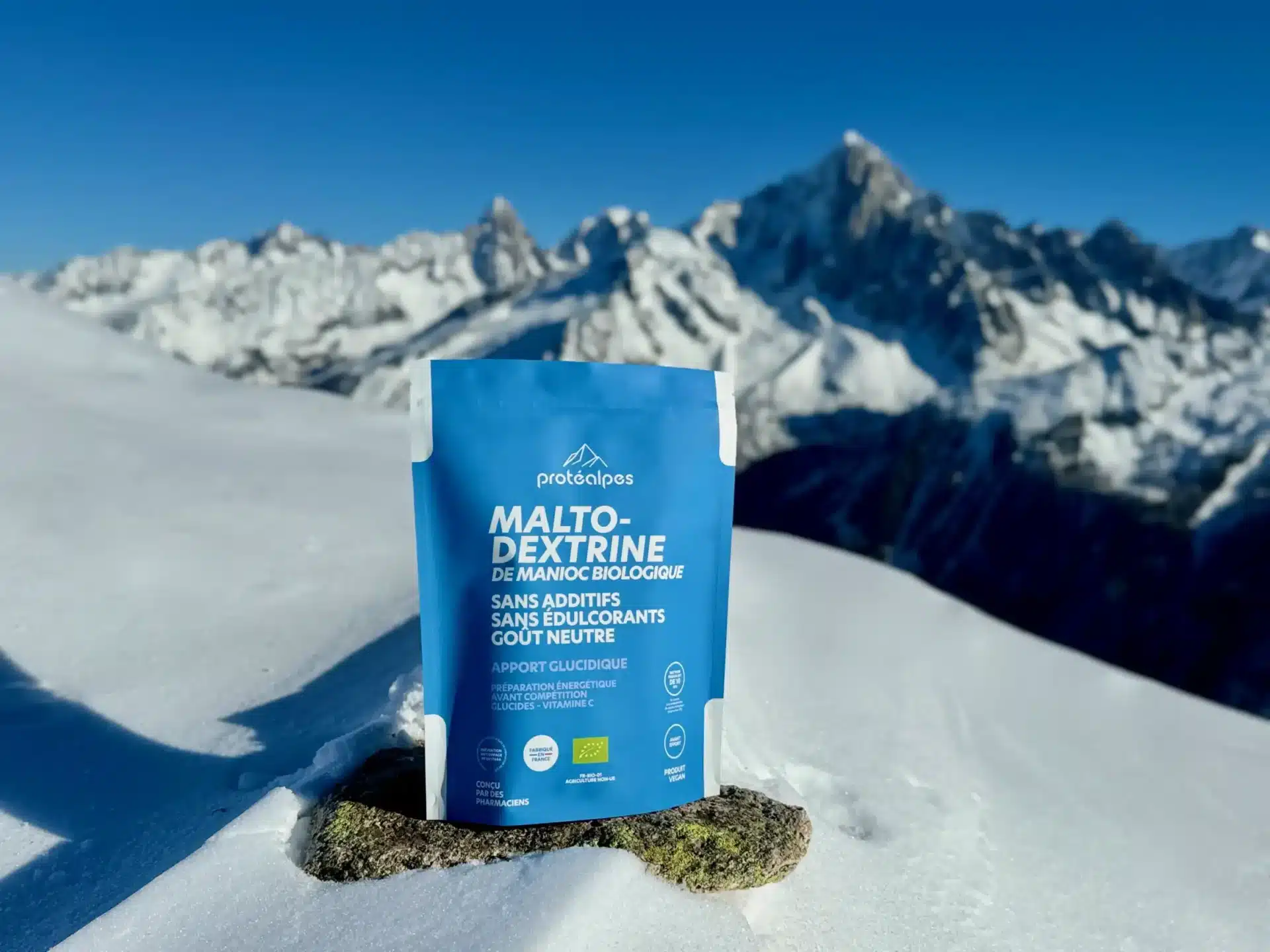 Sachet de maltodextrine Protéalpes sur neige, devant montagnes enneigées, soulignant nutrition sportive sans édulcorants ni additifs.