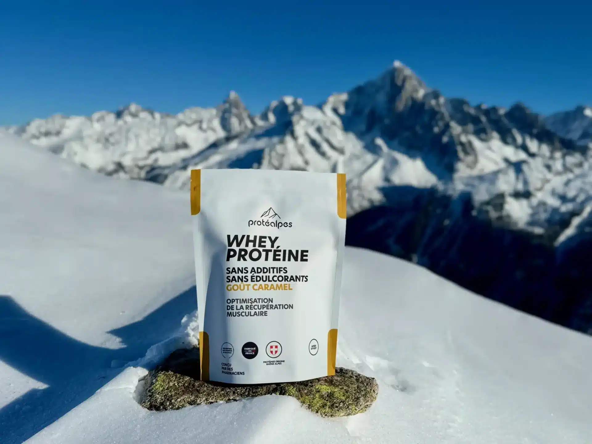 Sac de whey protéine Protéalpes goût caramel, sans édulcorants ni additifs, posé sur une neige avec montagnes enneigées en arrière-plan.