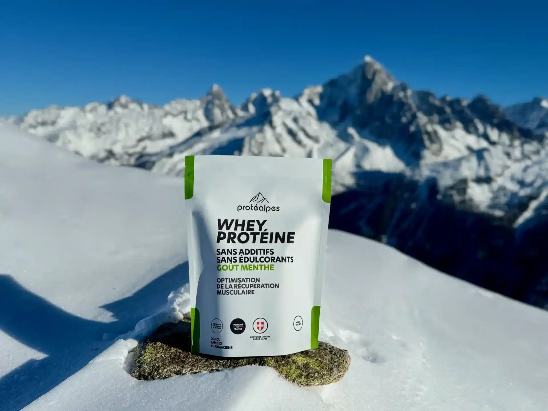 Sachet de protéine Protéalpes sur la neige, montagnes en arrière-plan. Sans édulcorants ni additifs, nutrition sportive pure.