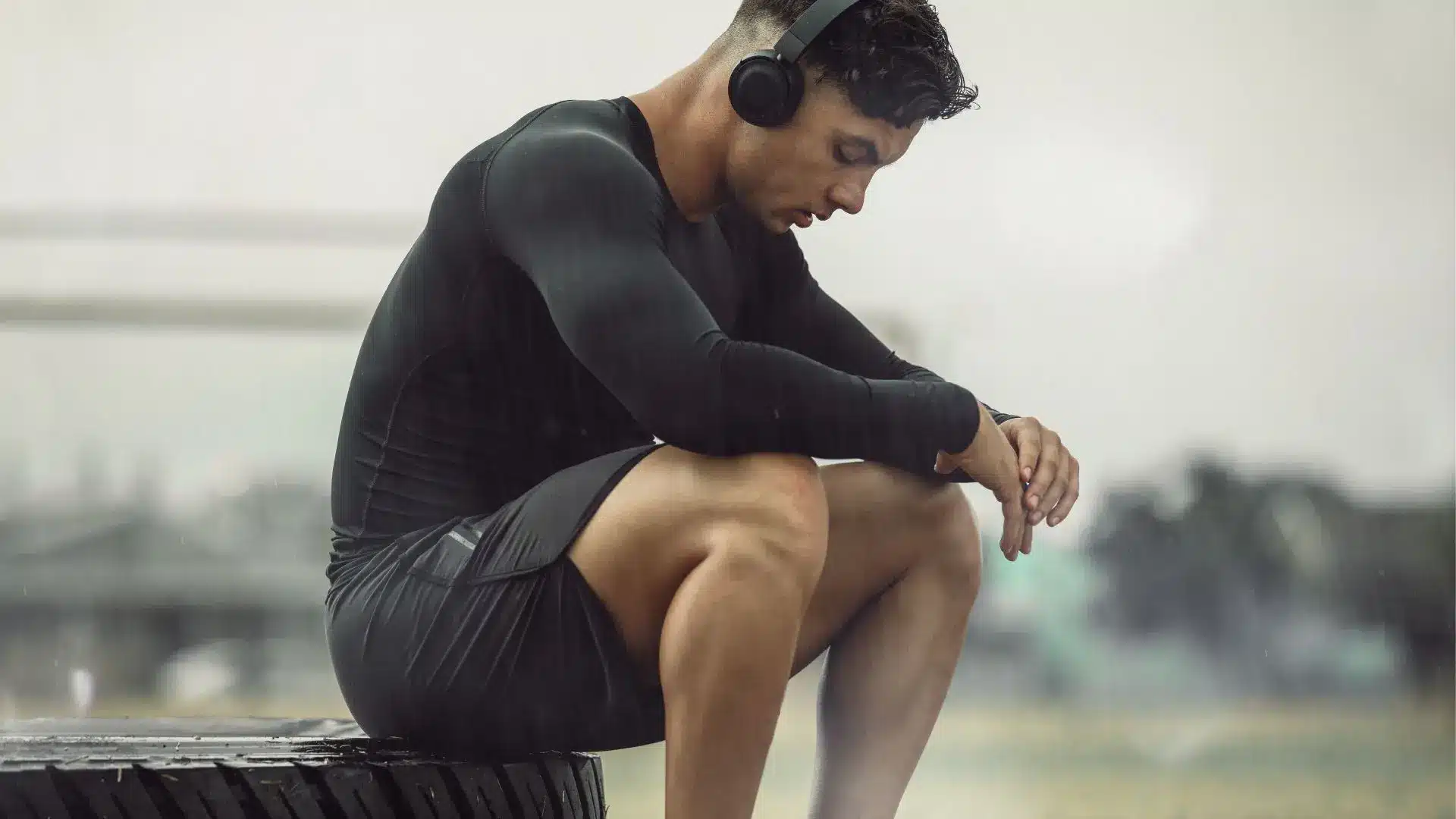 Homme en tenue sportive assis sur un pneu, écouteurs sur les oreilles, dans un moment de repos sous la pluie.